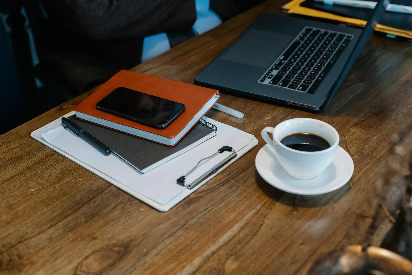 Mesa de madeira com celular sobre cadernos, prancheta, notebook e uma xícara de café