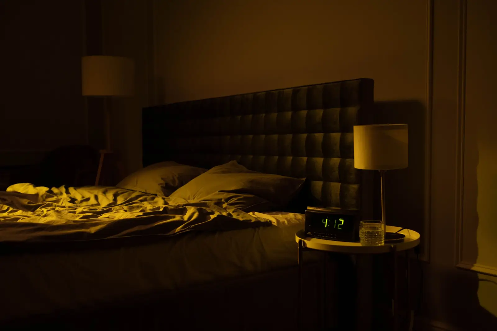 Quarto escuro à noite, com a cama desfeita e um relógio digital marcando 4h12 no criado-mudo
