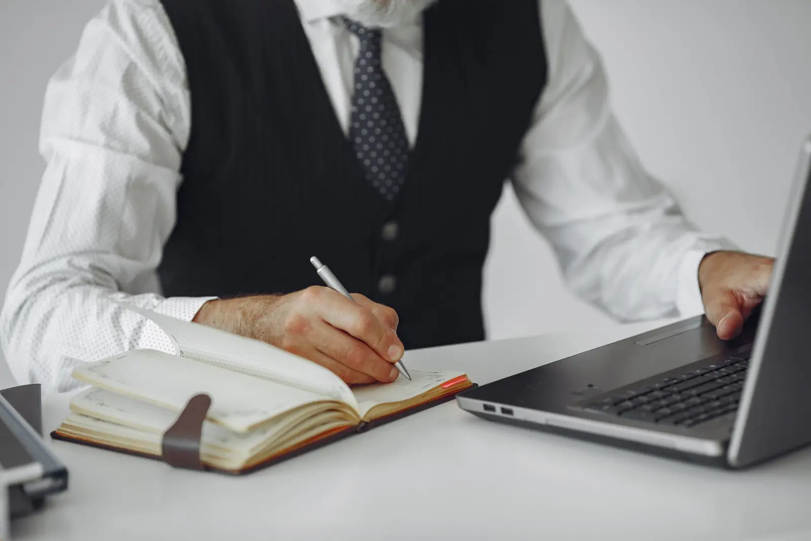 Homem de colete e camisa branca faz anotações em uma agenda, ao lado de um notebook, sobre uma mesa clara.