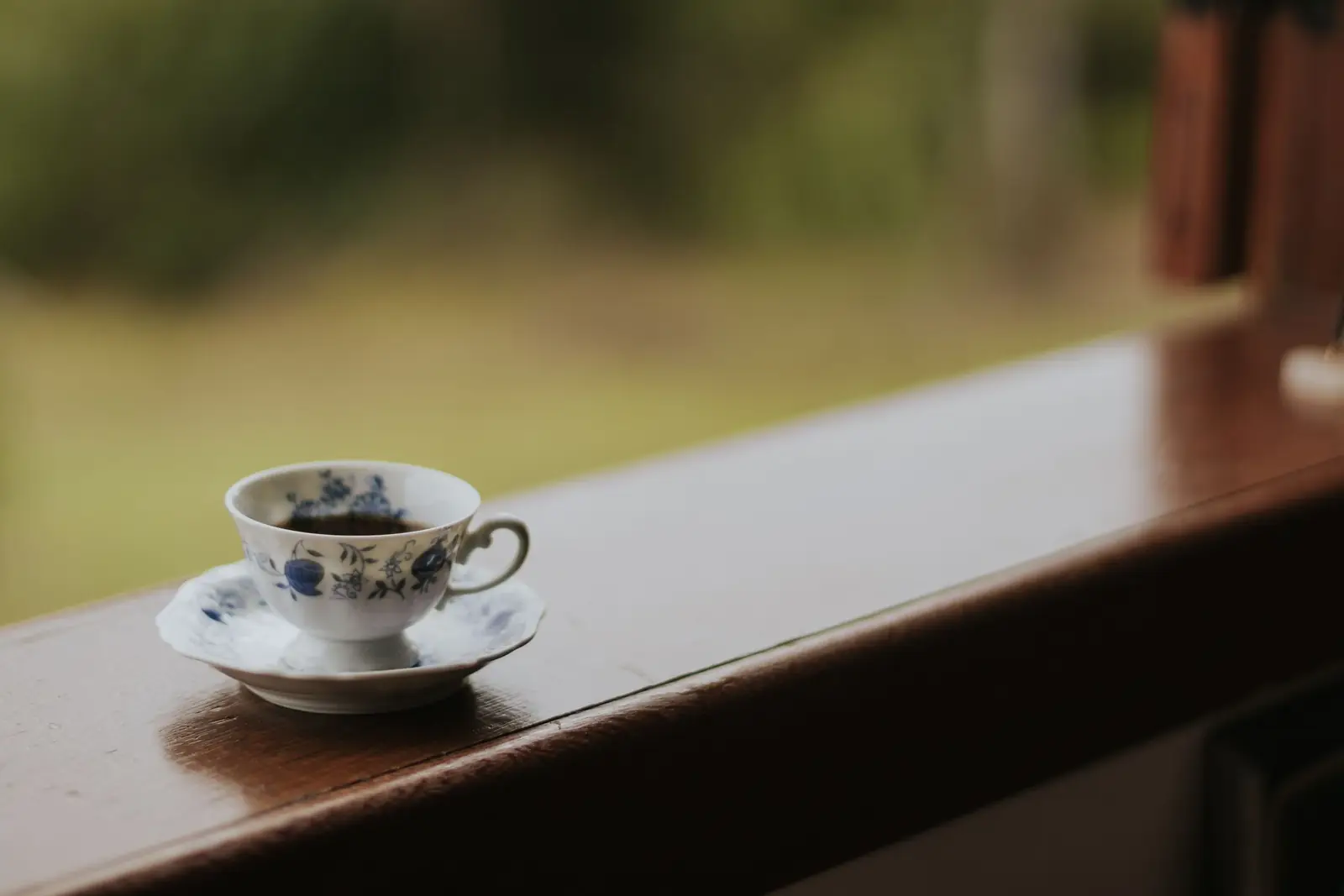 Xícara de porcelana com desenhos azuis sobre o parapeito de madeira de uma janela, com jardim desfocado ao fundo.