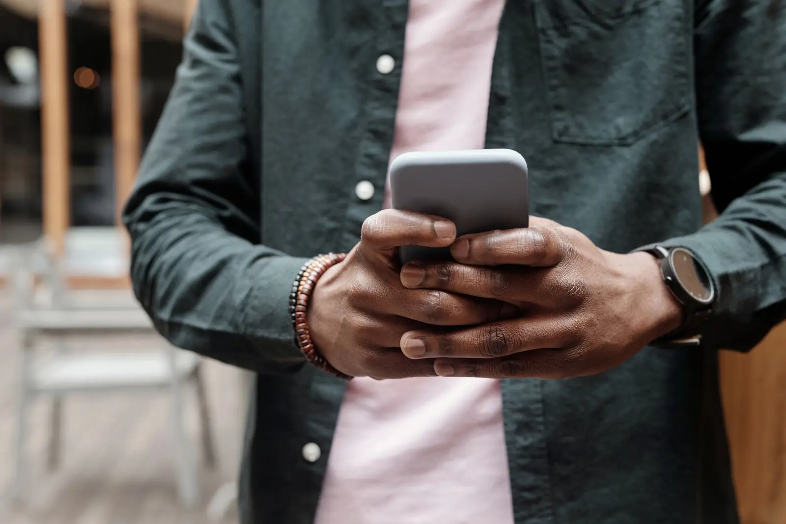 Mãos de um homem de camisa escura segurando o celular, com pulseira de contas e relógio no pulso