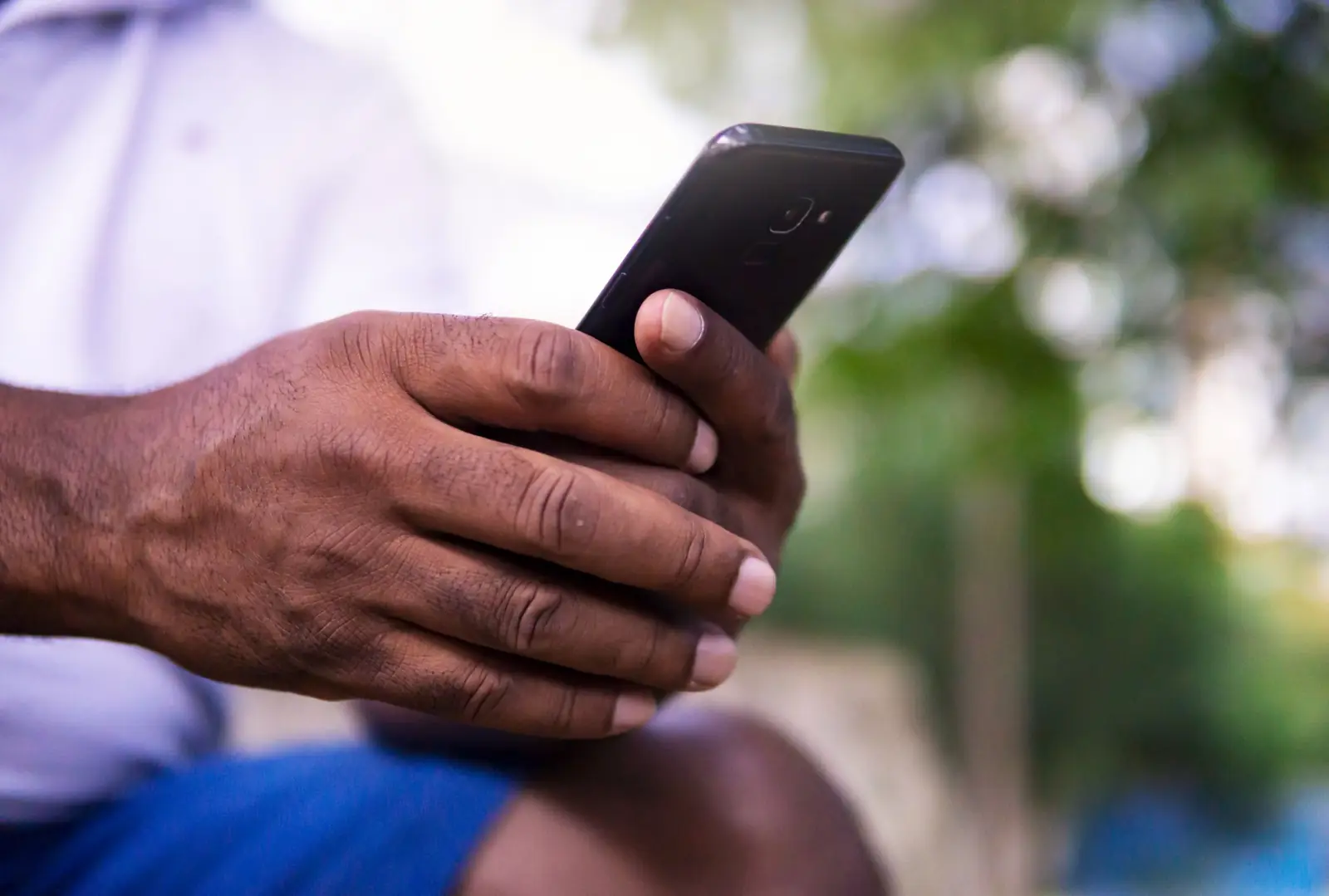 Mão de um homem segurando um celular preto ao ar livre, com árvores desfocadas ao fundo