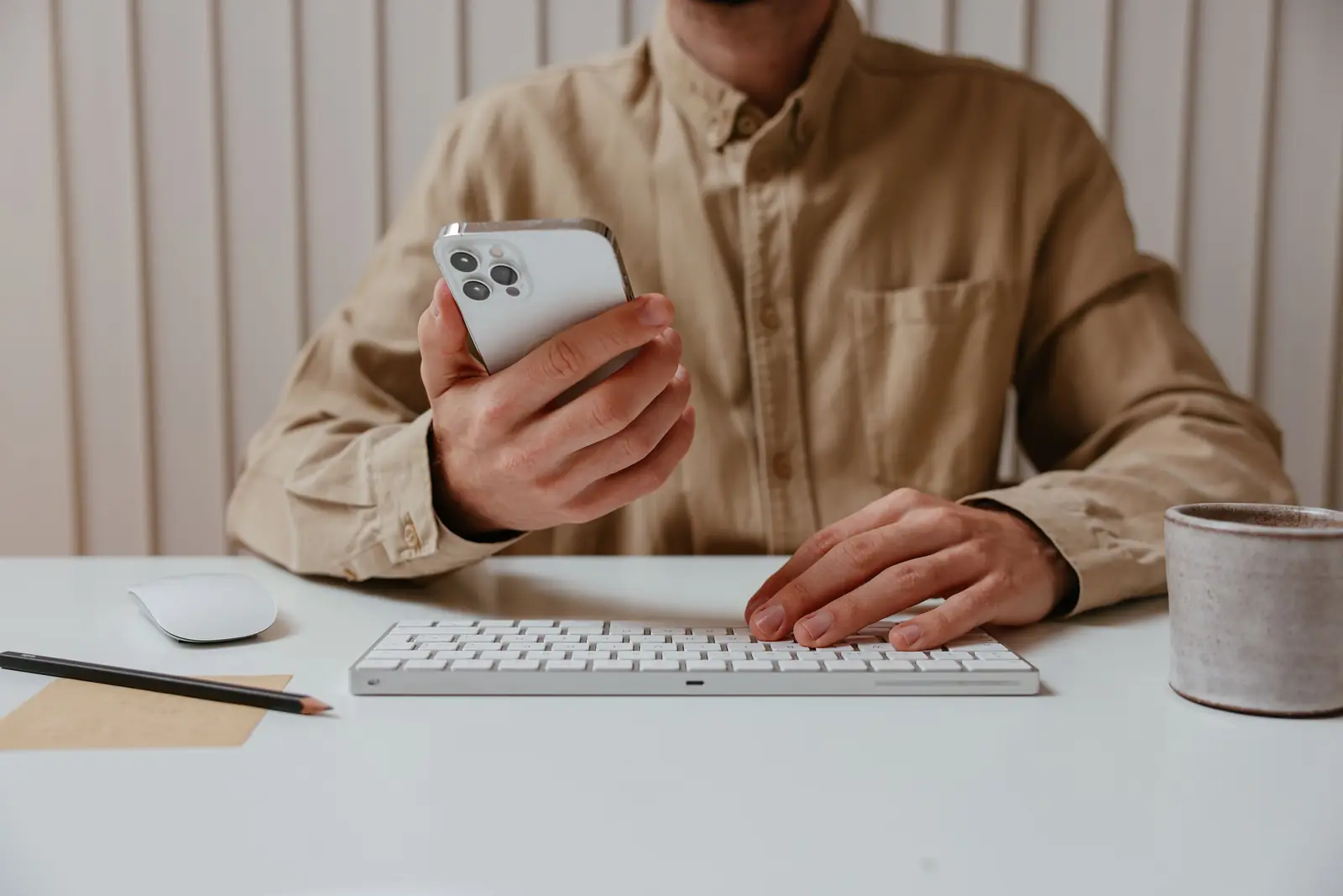 Homem de camisa bege segura o celular sentado à mesa, com teclado, caneca e lápis sobre o tampo branco.