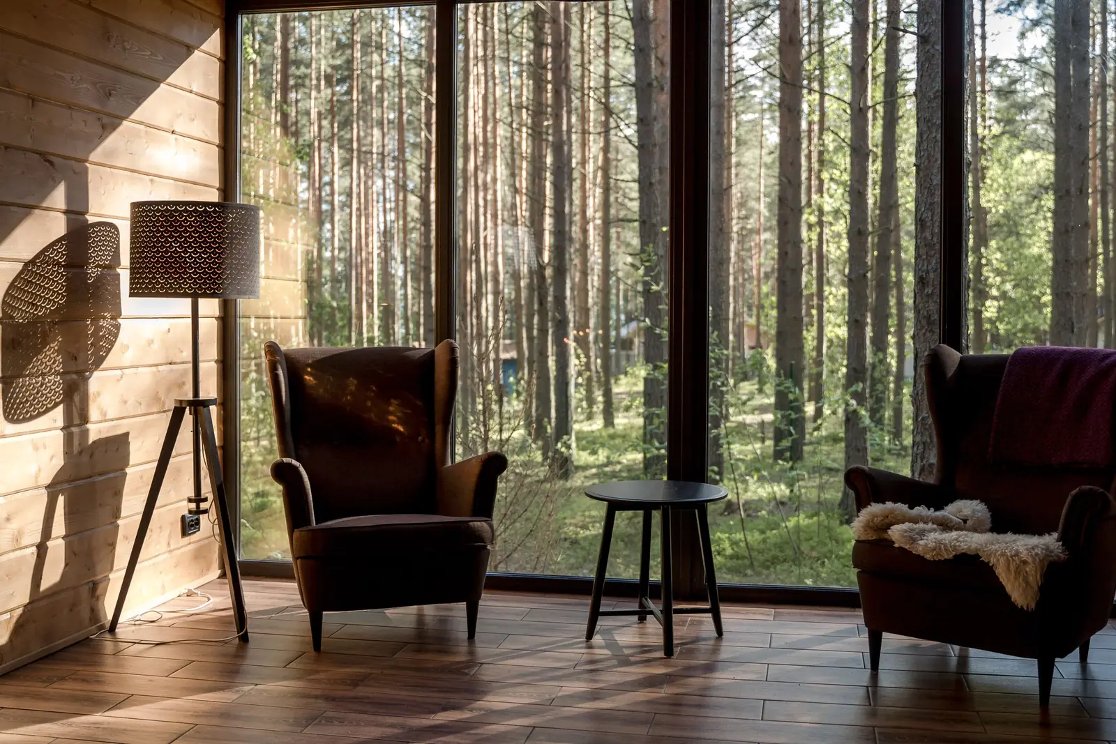Duas poltronas e uma mesinha diante de uma parede de vidro com vista para um bosque, sob luz natural