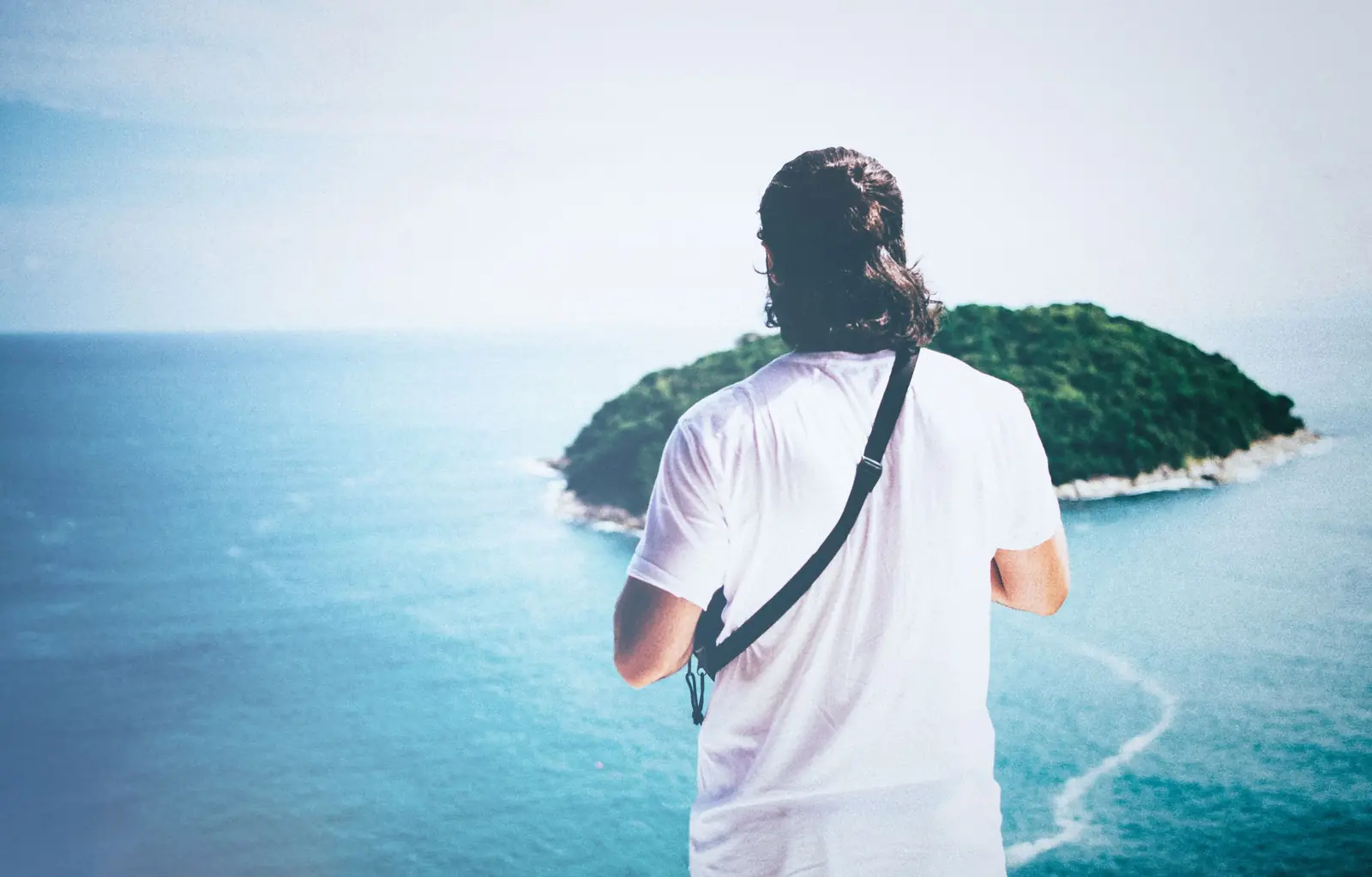 Homem de cabelo longo e camiseta branca, visto de costas, olha uma ilha verde no mar azul