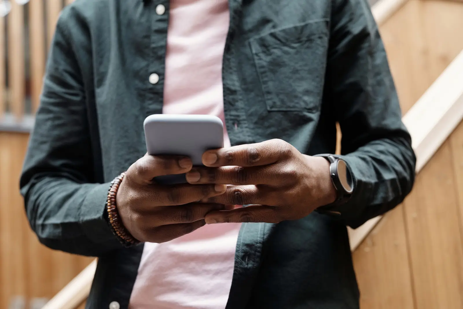 Homem de camisa verde-escura e relógio segura o celular com as duas mãos, diante de uma parede de madeira