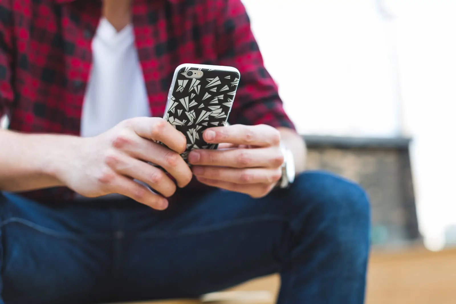 Homem sentado, de camisa xadrez vermelha e calça jeans, segura o celular com as duas mãos ao ar livre