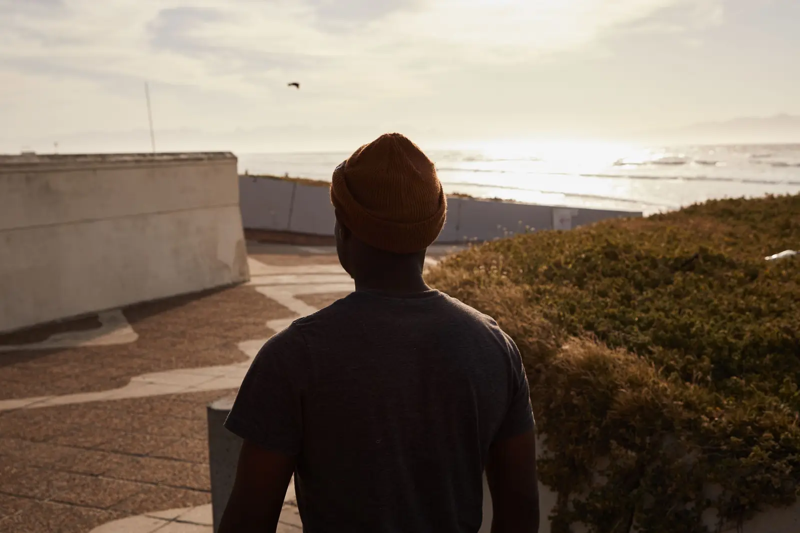 Homem de gorro, visto de costas, olha o mar de um calçadão, com o sol baixo no horizonte
