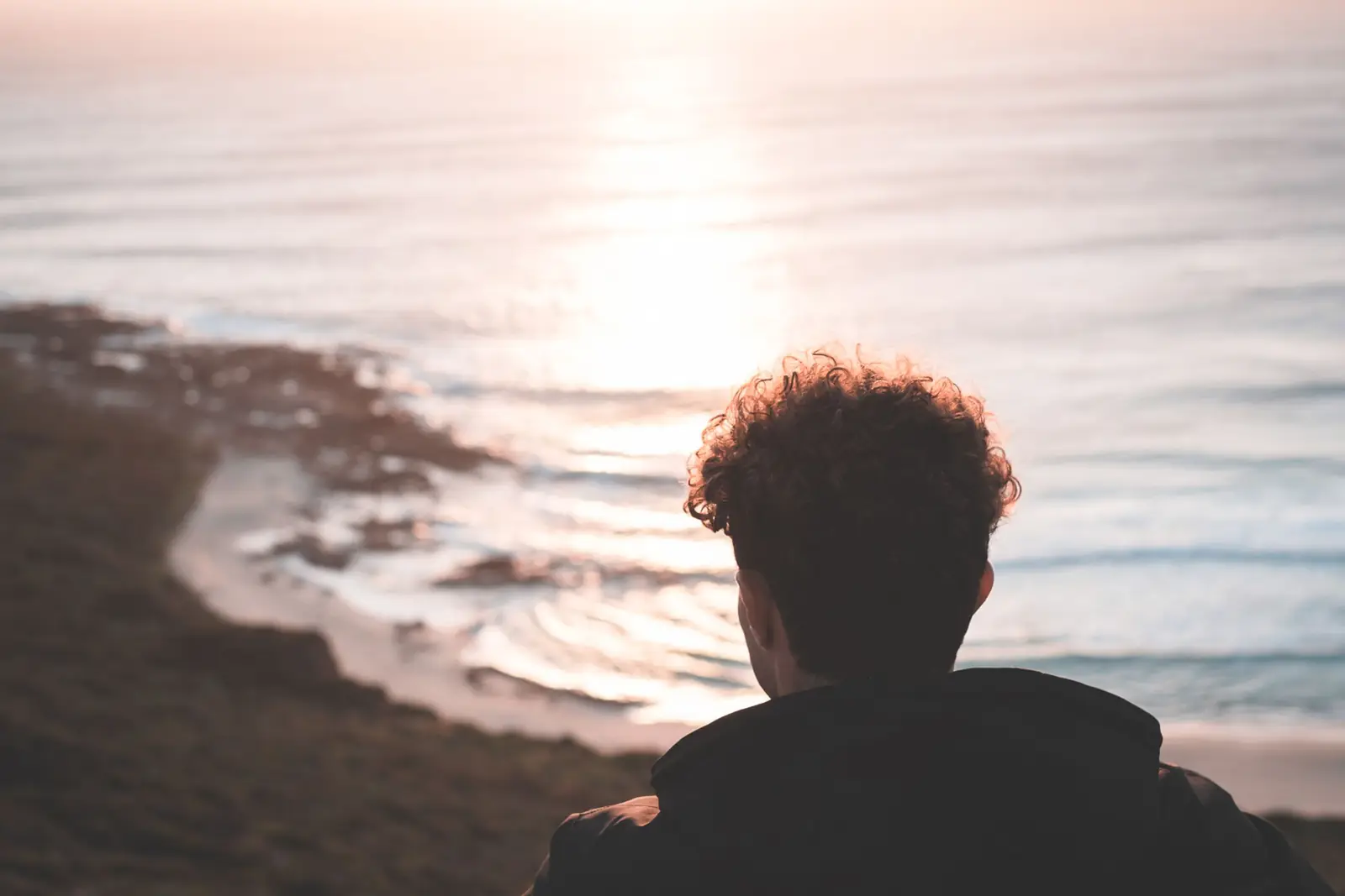 Homem de cabelo cacheado, visto de costas, observa o mar ao pôr do sol do alto de uma encosta
