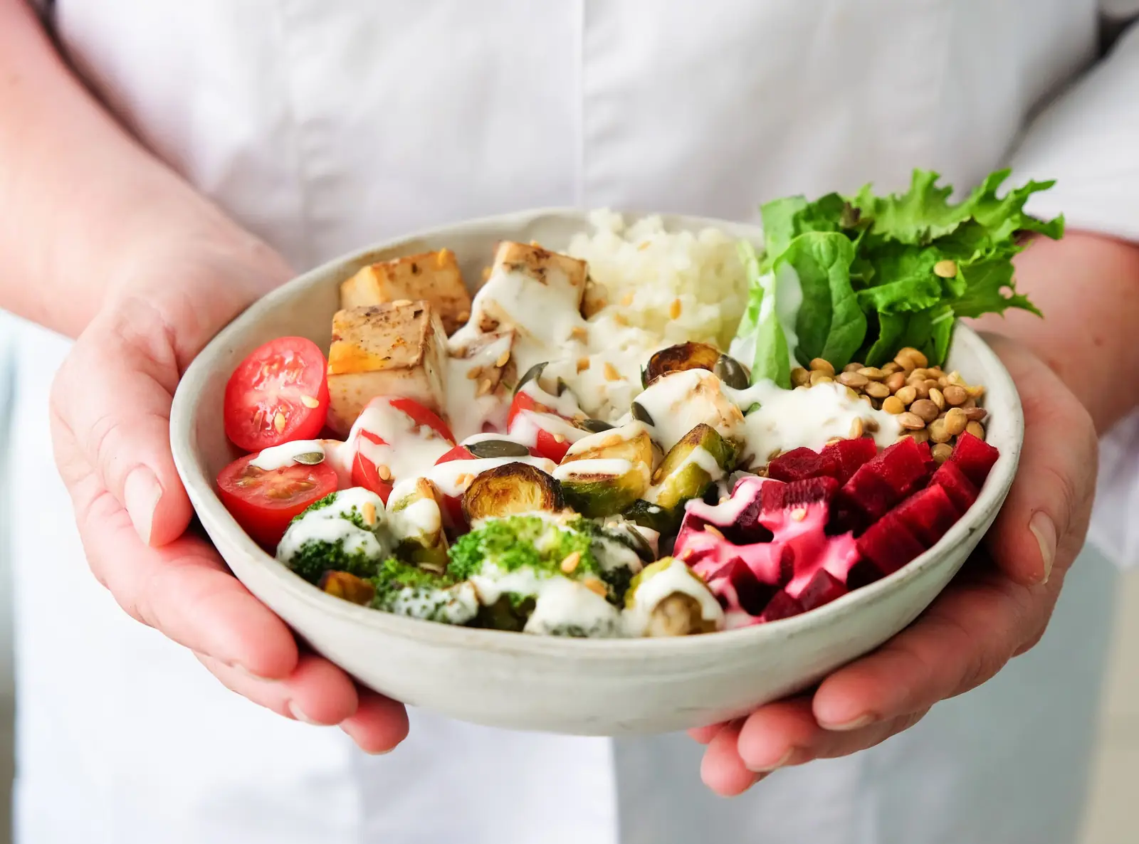Mãos seguram uma tigela com tofu, lentilhas, legumes assados, beterraba, tomate e folhas verdes.