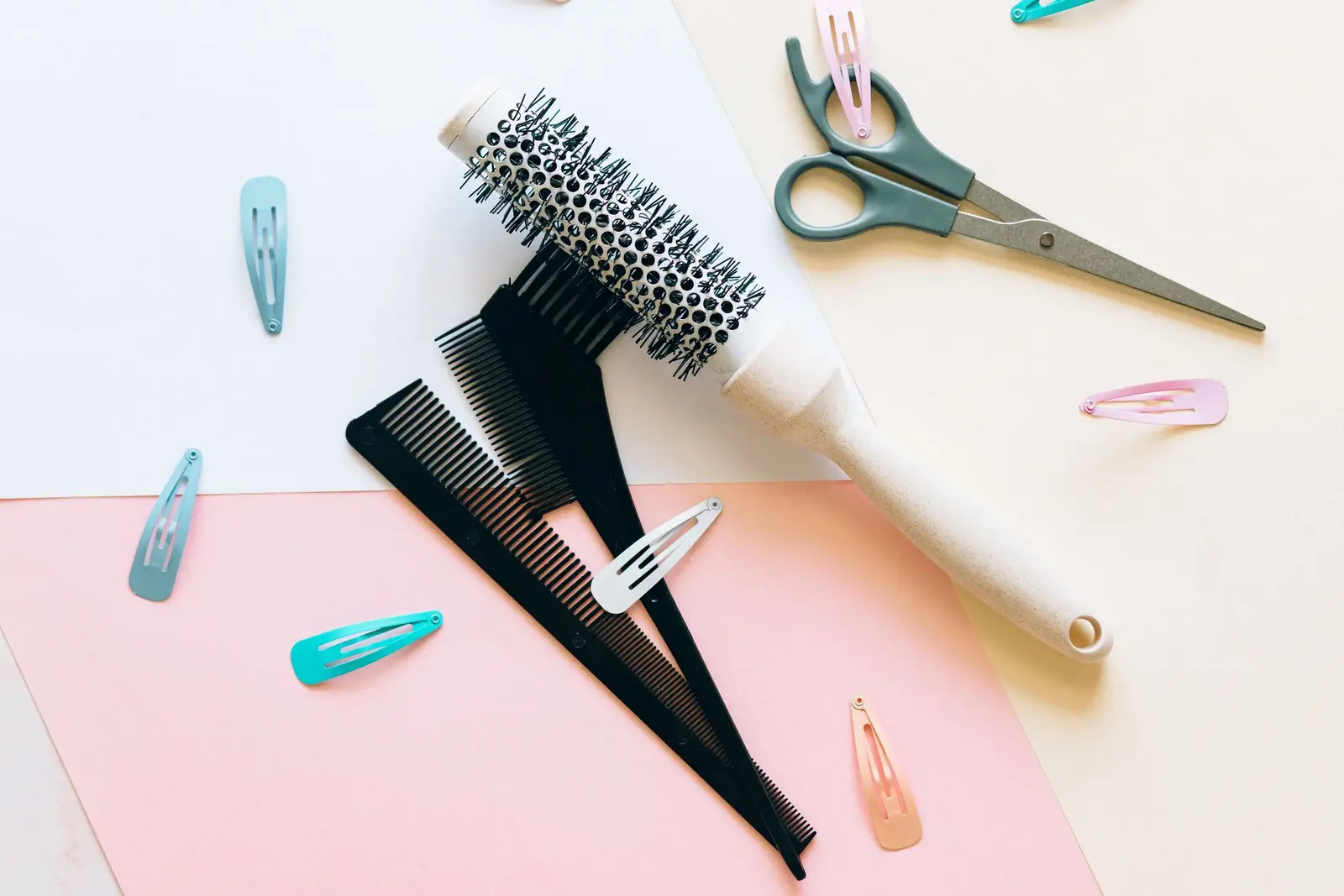 Escova redonda, pentes, tesoura e grampos de cabelo sobre papéis em tons de rosa e creme
