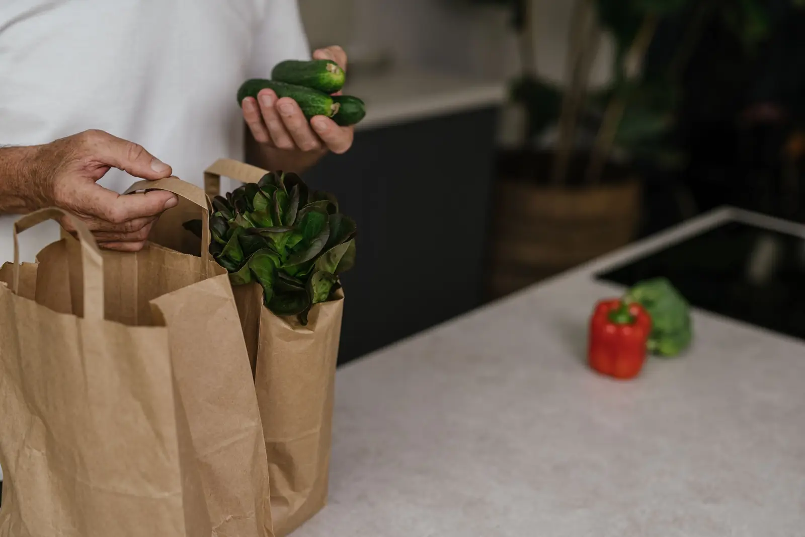 Mãos de um homem com pepinos, ao lado de sacolas de papel com alface, na bancada da cozinha.