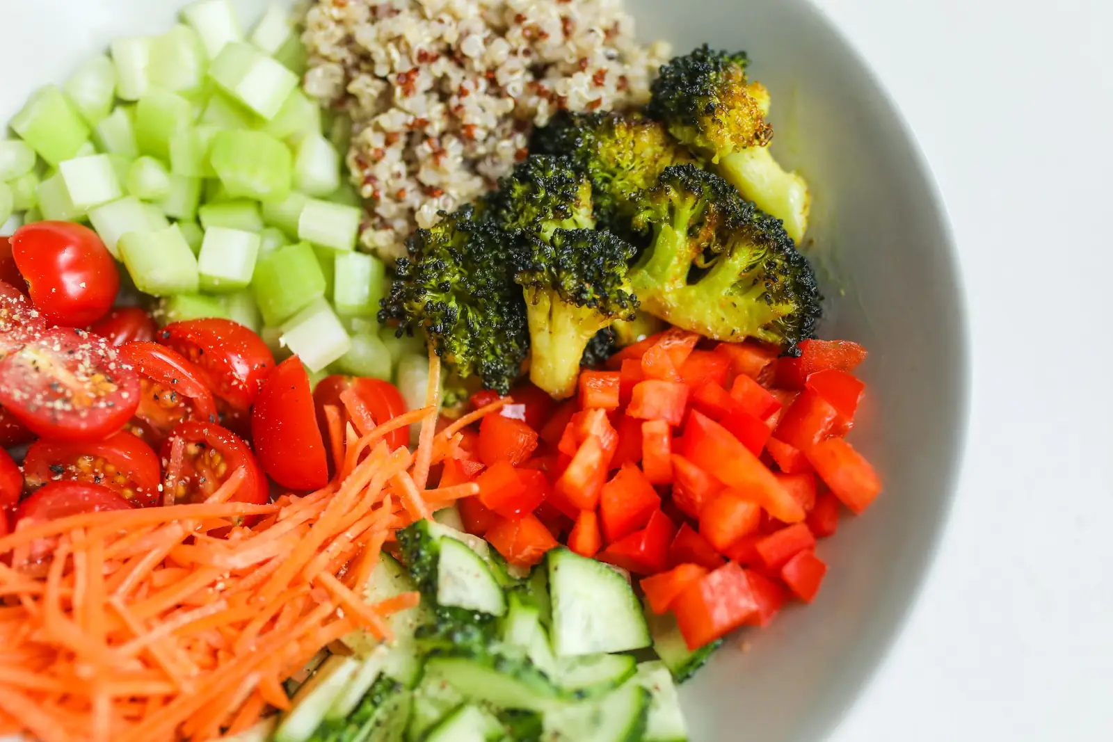 Tigela branca com quinoa, brócolis, tomate, cenoura ralada, pepino e pimentão picados, vista de perto