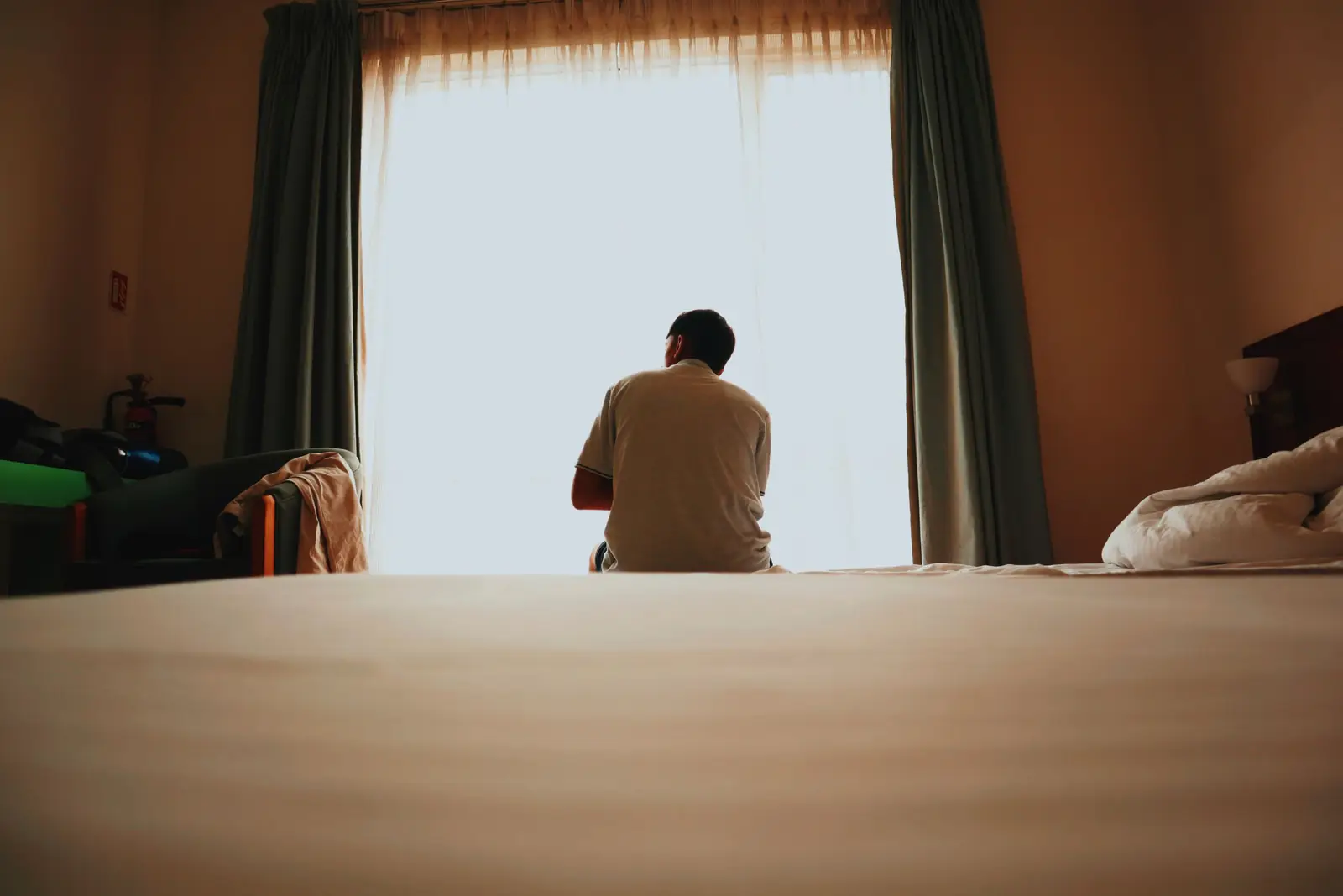 Homem visto de costas, sentado na beira da cama, olha para uma janela clara entre cortinas