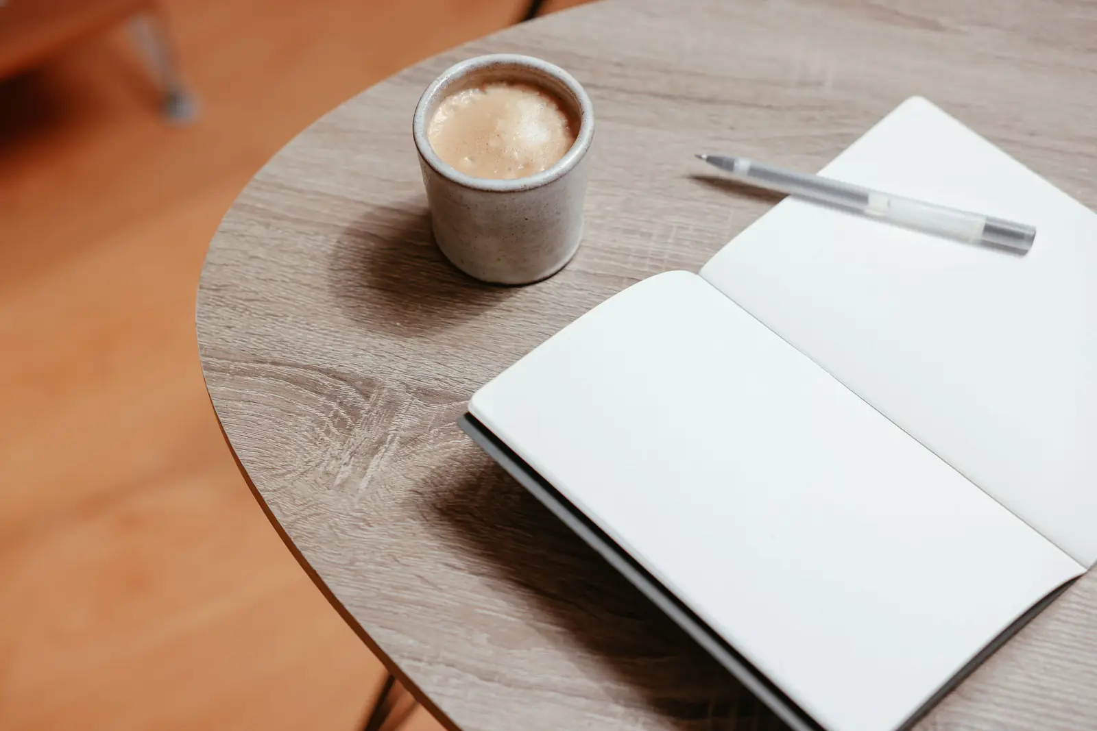 Caderno aberto com páginas em branco, caneta e copo de café sobre uma mesa redonda de madeira