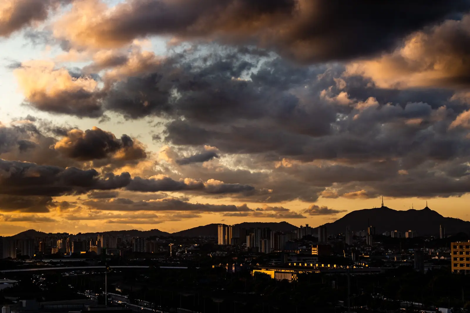 Nascer do sol sobre São Paulo, com prédios e montanhas sob nuvens iluminadas em tons dourados