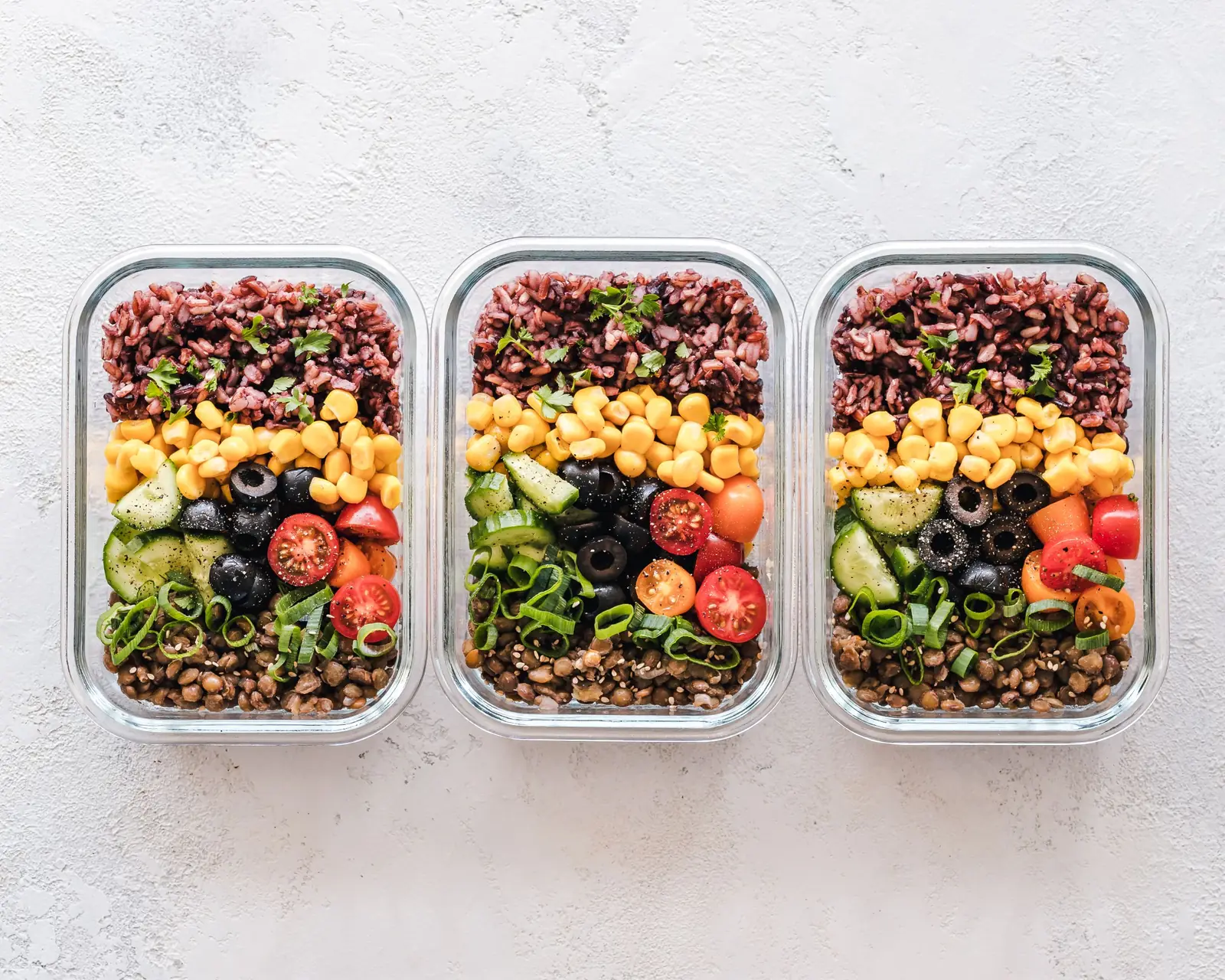 Três potes de vidro com refeições prontas de arroz, milho, legumes e lentilha sobre uma bancada clara