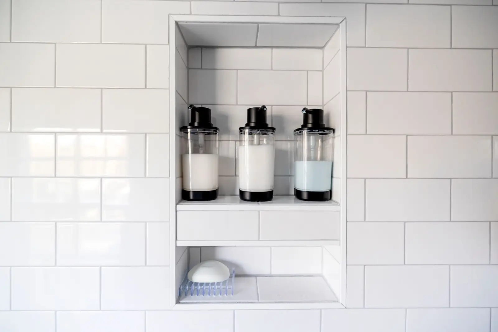 Nicho de banheiro com azulejos brancos e três frascos dispensadores de xampu e condicionador