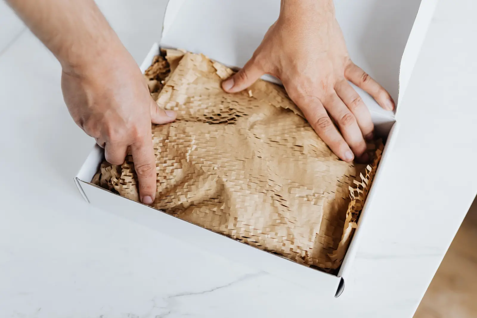 Mãos de um homem abrindo uma caixa branca forrada com papel kraft sobre uma mesa clara
