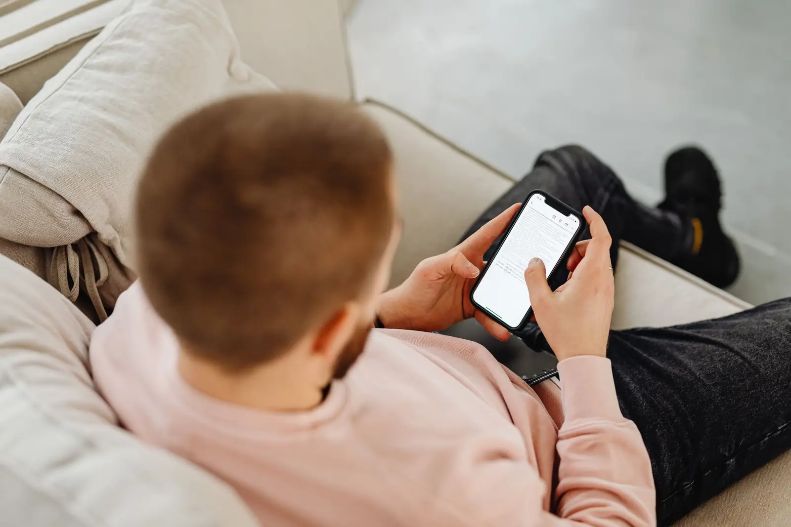 Homem visto de costas, sentado em um sofá claro, usa o celular com as duas mãos