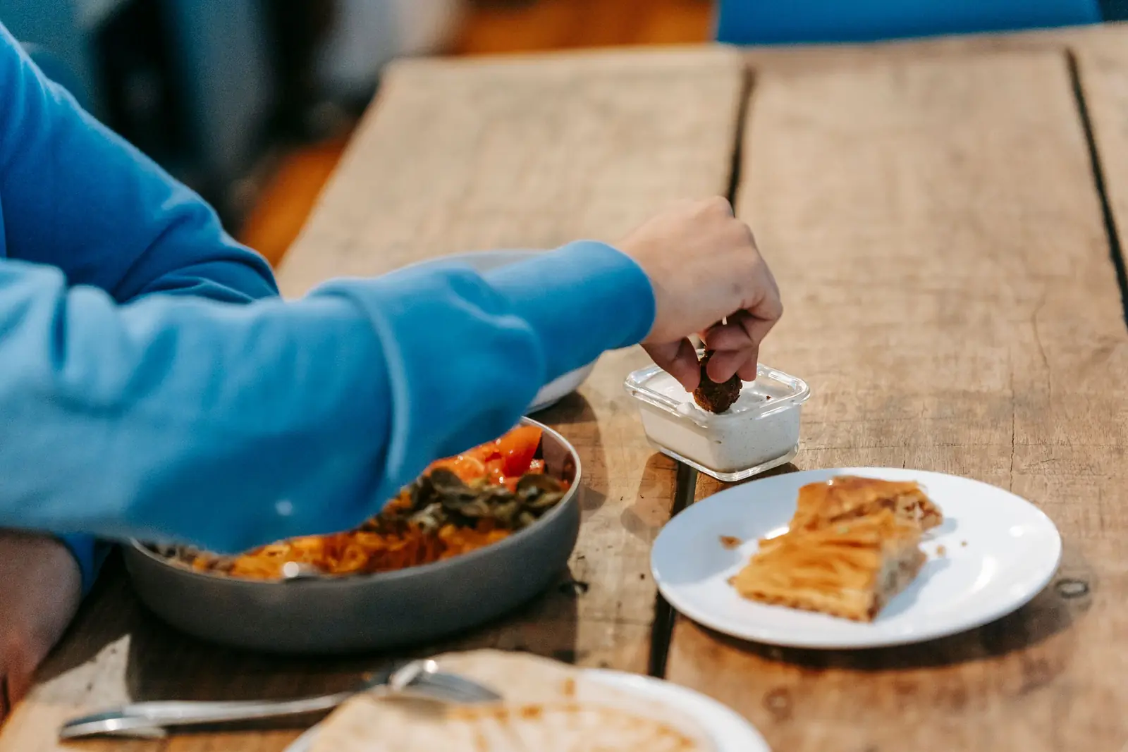 Homem de blusa azul faz uma refeição em uma mesa de madeira, com prato de torta e travessa de comida.